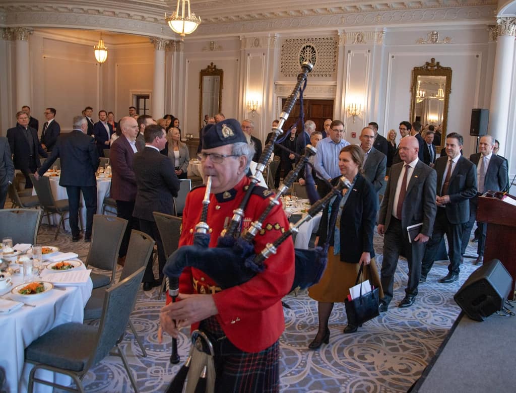 A piper in a red tunic leads guests through a crowded luncheon